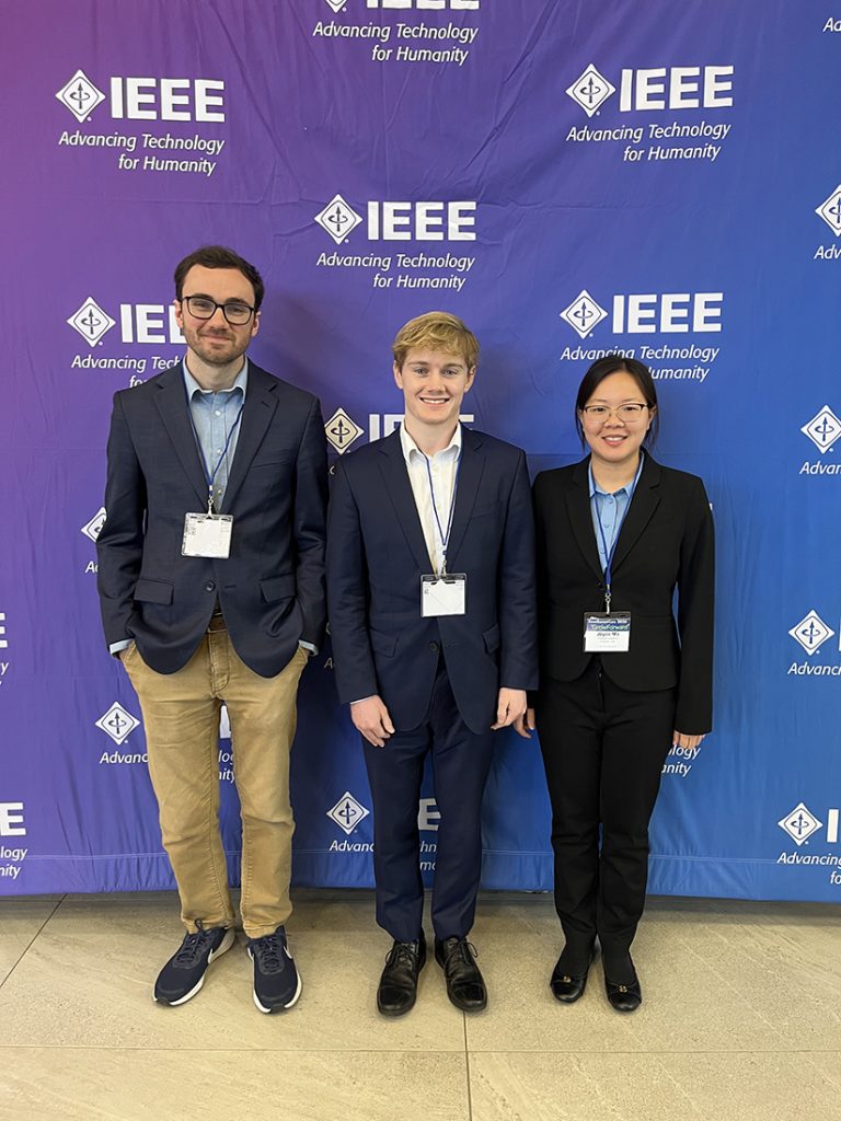 3 students standing in front of a IEE logo background